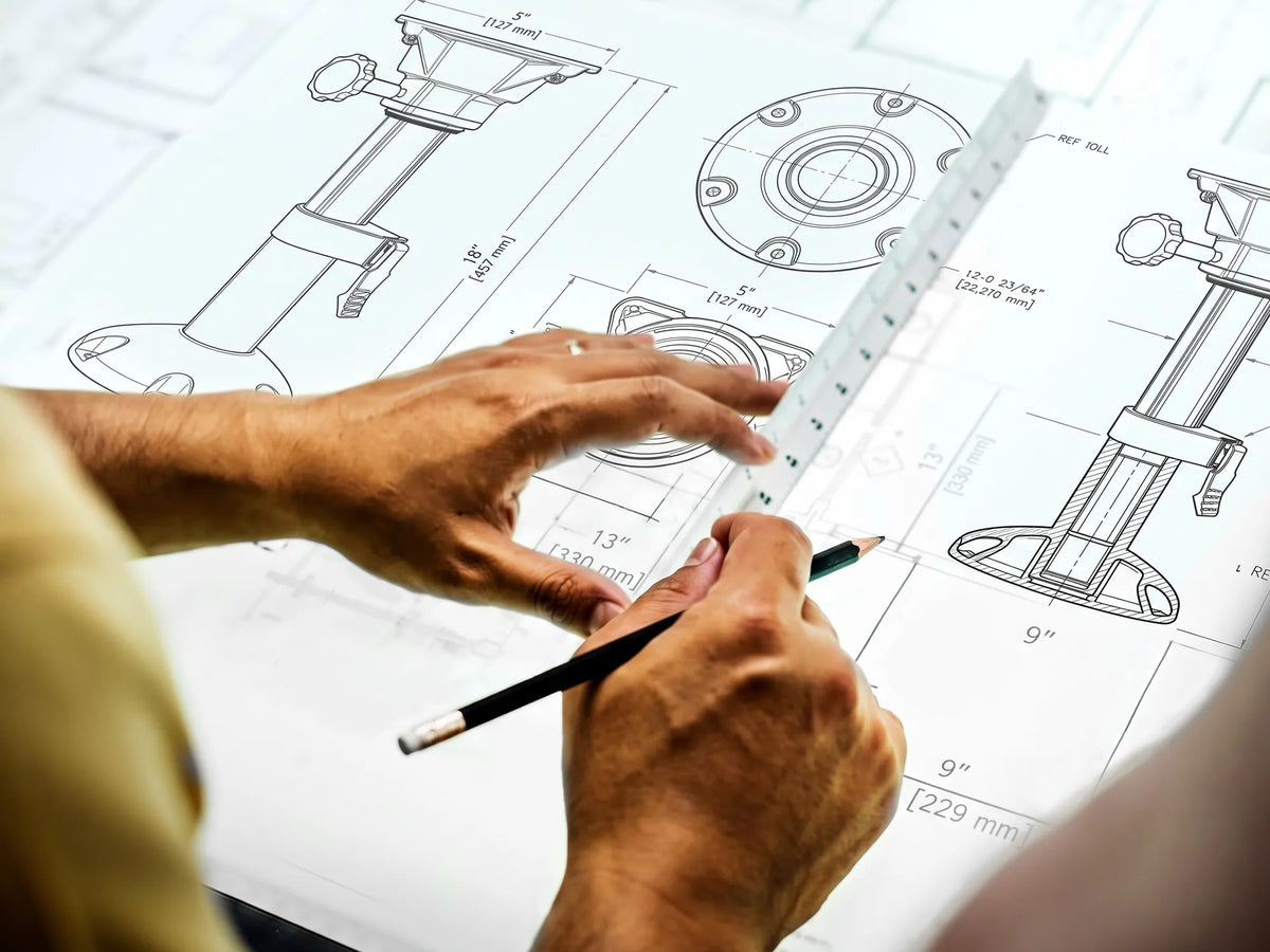 Hands reviewing boat seat pedestal technical drawings with a ruler and pencil on a drafting table.