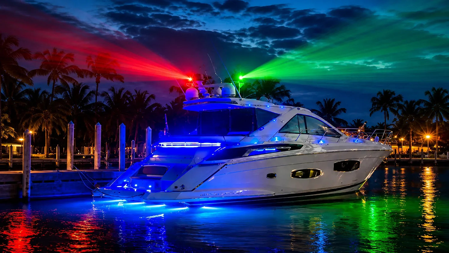 Luxury yacht docked at a marina at night with red and green navigation lights reflecting on the water.