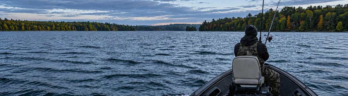 Angler seated on a gray fishing boat seat with two rods facing a wooded lake shoreline.