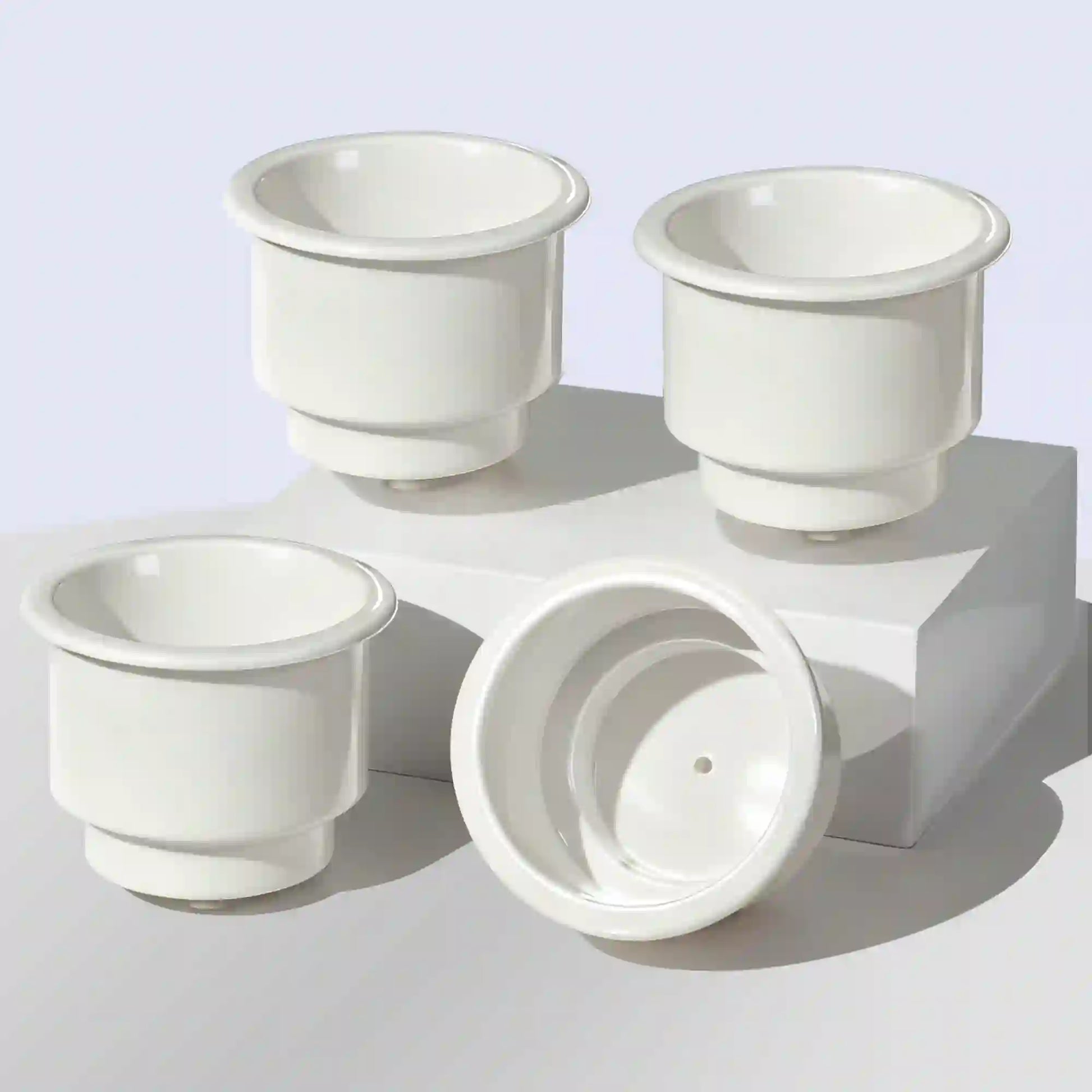 Four Marinexplore white ABS plastic boat cup holder inserts arranged on a white cube display surface, with three units showing the smooth round interior opening from above and one unit flipped upside down to reveal the centered drain hole on the bottom, photographed against a light blue background.