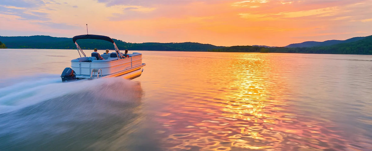 Pontoon boat carrying passengers across a calm lake beneath a colorful sunset sky reflected on the water.