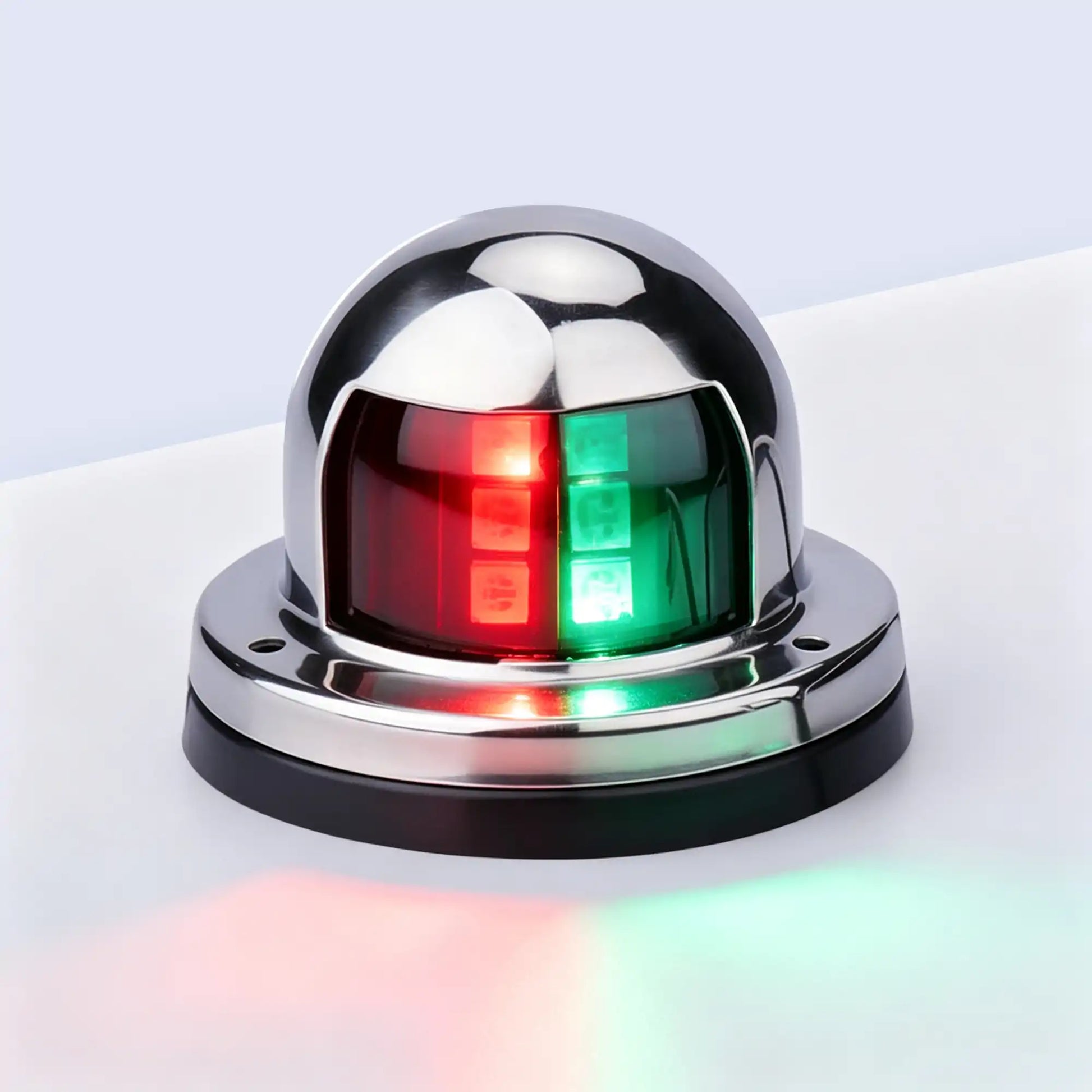 Marinexplore chrome-plated stainless steel boat bow navigation light with a rounded dome housing and black rubber base, showing a bicolor lens glowing red on the left and green on the right, photographed at a three-quarter angle against a light gray studio background with soft light reflections spilling onto the surface below.